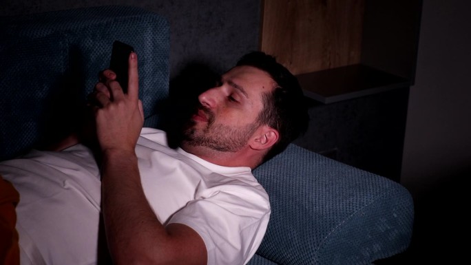 晚上坐在家里舒适的客厅沙发上玩智能手机的帅哥。