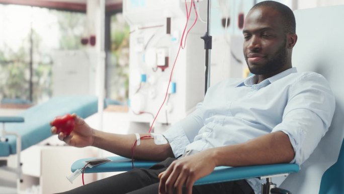 黑人商人为有需要的人献血。一名非洲男子挤压心形红球，将血液通过管道泵入袋子，并在镜头前展示，作为支持