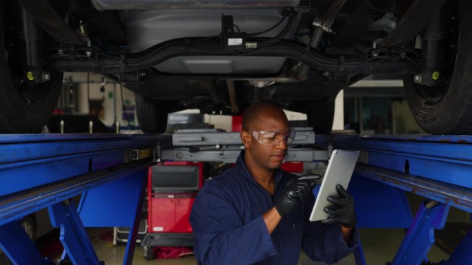 在一家汽车修理店，黑色机械师一边检查平板电脑上的清单，一边检查汽车底部