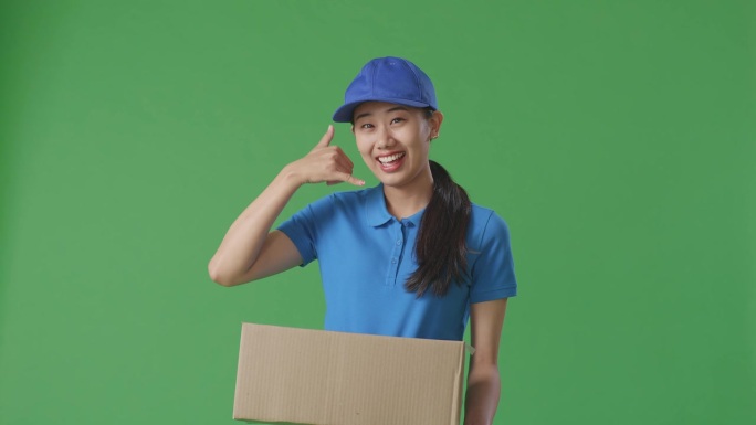 蓝色制服的亚洲女快递员在绿屏背景下递纸盒时，做着叫我的手势和微笑