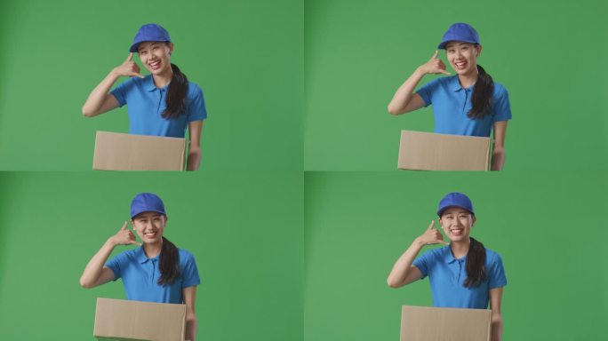 蓝色制服的亚洲女快递员在绿屏背景下递纸盒时，做着叫我的手势和微笑