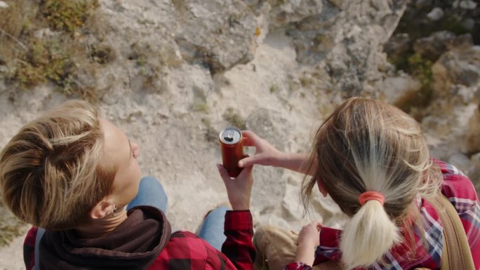 女性朋友徒步旅行者喝着锡罐里的饮料，一边放松旅行，一边坐在岩石海岸的边缘山上，欣赏着令人惊叹的景色