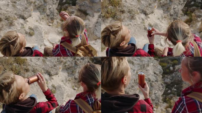女性朋友徒步旅行者喝着锡罐里的饮料，一边放松旅行，一边坐在岩石海岸的边缘山上，欣赏着令人惊叹的景色