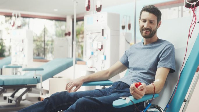 白人男子在光明医院为有需要的人献血。男性献血者挤压心形红球，将血液通过管道泵入袋中。为烧伤病人捐款。