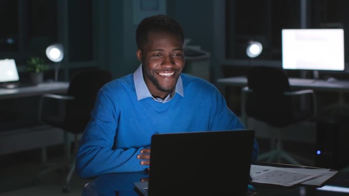 快乐的非裔美国商人用笔记本电脑开视频会议，在漆黑的空办公室里工作到很晚