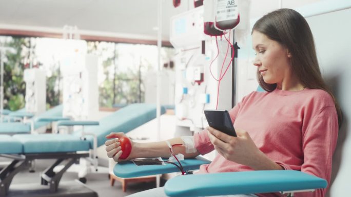 白人妇女在医院为有需要的人献血。女献血者挤压心形红球泵血，用智能手机在线聊天。器官移植病人的捐赠。