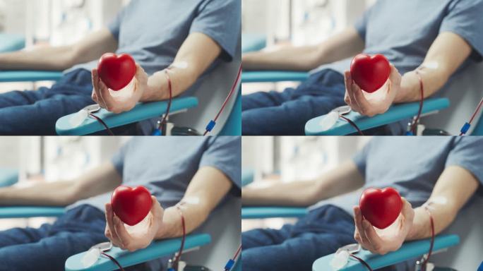 男性献血者的手部特写与置管。一名白人男子挤压心形红球，将血液通过管道泵入袋子，并在镜头前展示，以示支