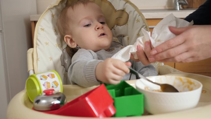 母亲用纸巾擦拭她脏兮兮的小男孩坐在高脚椅上喝粥的画像。育儿理念，健康营养和婴儿喂养