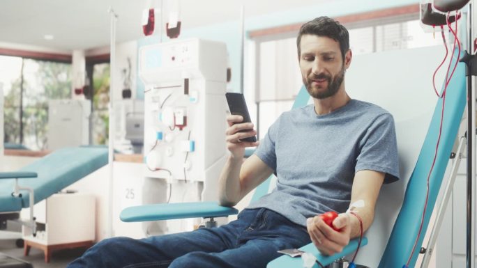 白人男子在医院为有需要的人献血。男性献血者挤压心形红球泵血，用智能手机在线聊天。器官移植病人的捐赠。