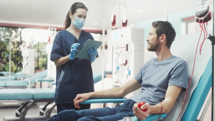 白人男子在医院为有需要的人献血。拿着药片的女护士过来检查捐赠者的进展和健康状况。捐献给需要输血的病人