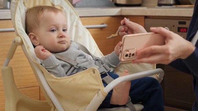 小男孩坐在高脚椅上一边看动画片一边喝粥的照片。育儿理念，健康营养和婴儿喂养