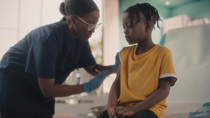一个年轻的非裔美国男孩坐在光明医院的椅子上接种小儿麻痹症疫苗。黑人女护士正在打针。职业女性与焦虑的孩