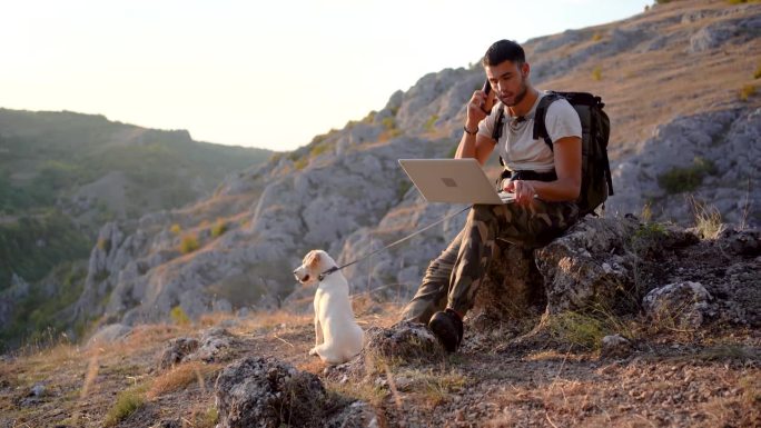 年轻人和他的狗在山上徒步旅行后，在大自然中打电话和使用笔记本电脑