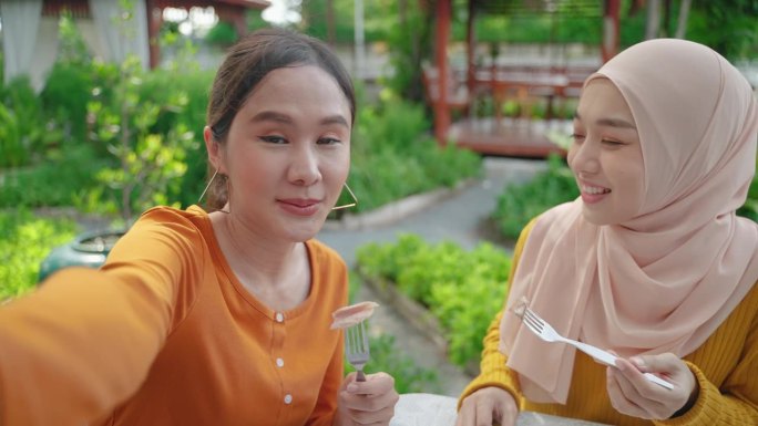 一名年轻的东南亚女子和她的穆斯林朋友在餐厅吃饭时在社交媒体上自拍，向朋友们展示食物。