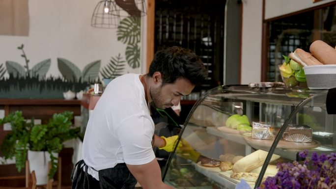 一名印度男子经营着一家小型家庭咖啡店。帮妻子清洗放蛋糕的玻璃柜。准备好每天早上开门营业。随时准备接待