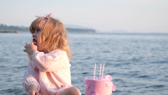 一个小女孩穿着粉红色的衣服，坐在那里吃着粉红色的蛋糕，拿着三支蜡烛和一支笔，背景是美丽的大海，晚上阳