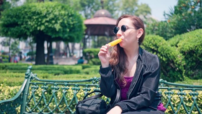 夏日周末，一名年轻女子戴着墨镜坐在墨西哥城科约阿坎公园的长椅上，一边吃着美味的橙色水果冰淇淋