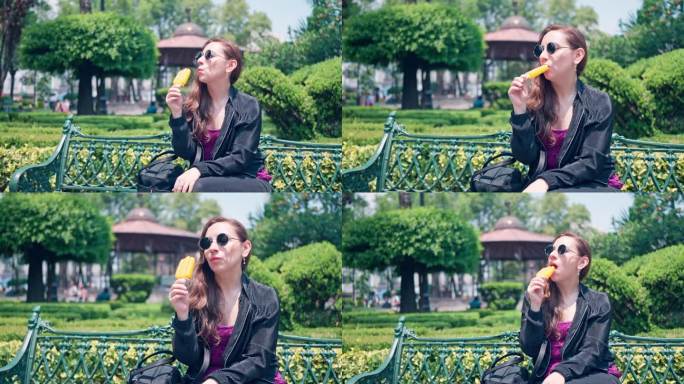 夏日周末，一名年轻女子戴着墨镜坐在墨西哥城科约阿坎公园的长椅上，一边吃着美味的橙色水果冰淇淋