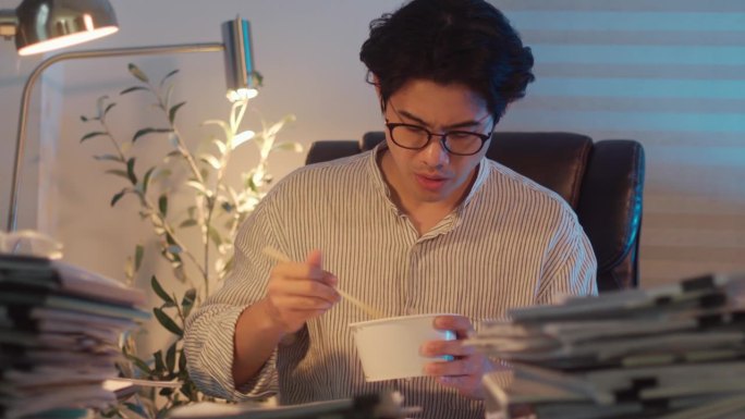 一个年轻的商人一边在家里用笔记本电脑工作一边吃方便面的4k视频片段。晚上加班