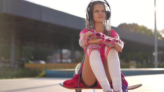 一个放松的少女坐在滑板上，戴着耳机听音乐。白人少年在城市的日出阳光下享受业余爱好。缓慢的运动。