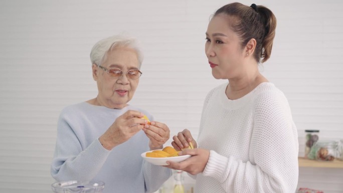 一位年长的亚洲妇女和中年的女儿在厨房里吃桔子。幸福的夫妇从橙子中摄取维生素C，在假期中使健康免于感染