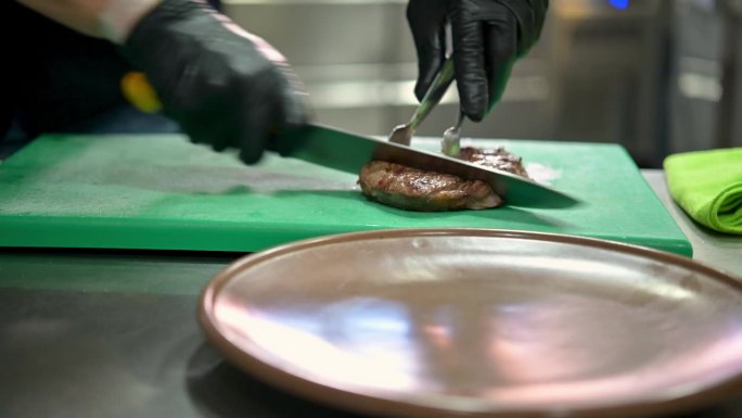 实时手持拍摄作物匿名厨师戴着黑色手套用刀切肉饼，然后把肉块夹在盘子上，而在餐厅厨房烹饪