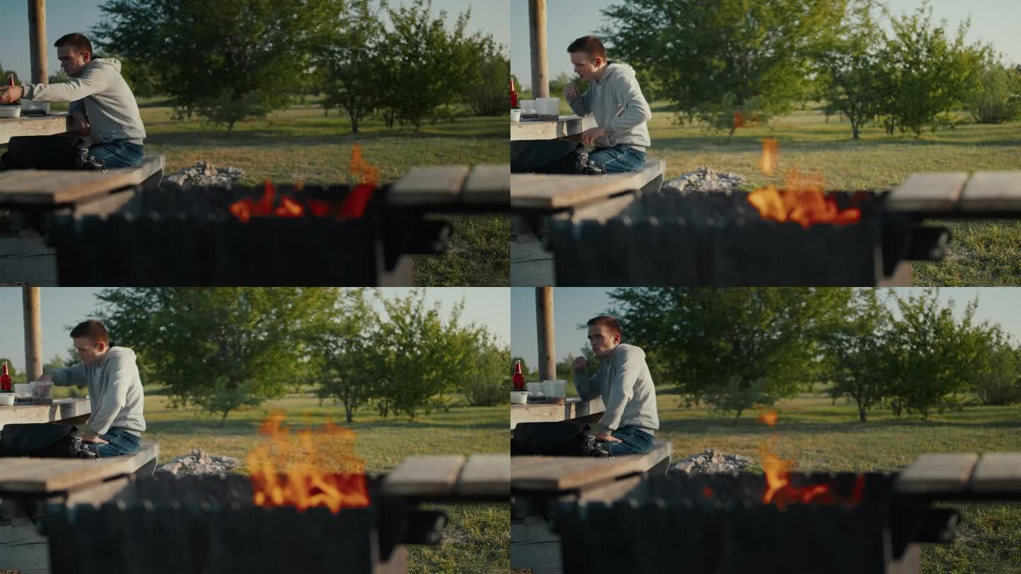 一个人坐在桌子上细细品味着他的食物，而他面前的烤架则从熊熊的火焰中散发出温暖