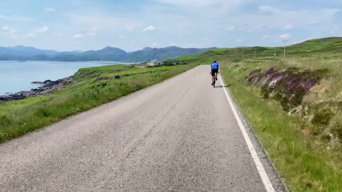 一名男子骑着自行车沿着风景如画的海岸公路驶向苏格兰的绿色山脉