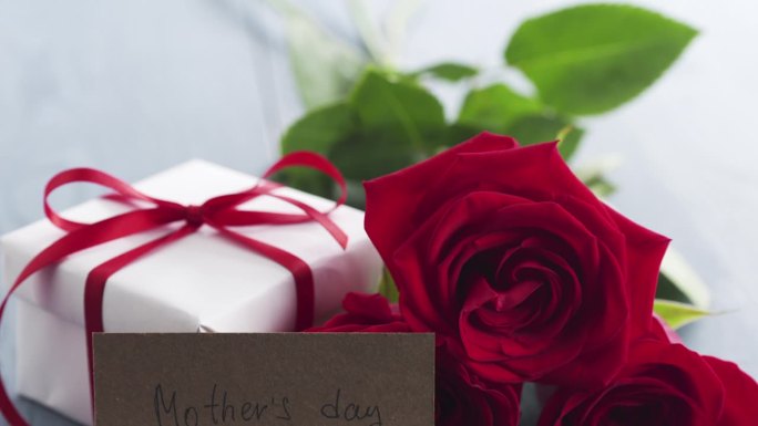三朵红玫瑰，礼盒和印有母亲节字样的纸卡，放在蓝色的木桌盘上