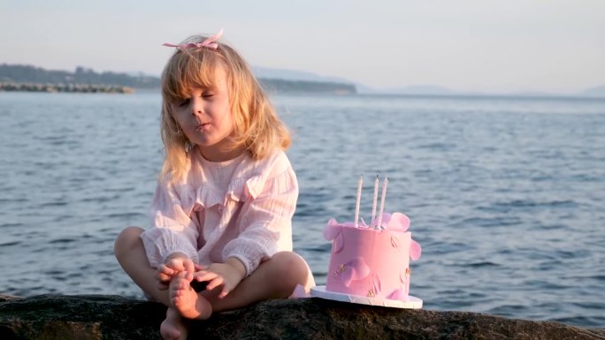 小女孩在粉红色的裙子手指看着咀嚼美味的粉红色蛋糕与她的生日3年海洋海洋海滩美丽欢乐爱家庭庆祝