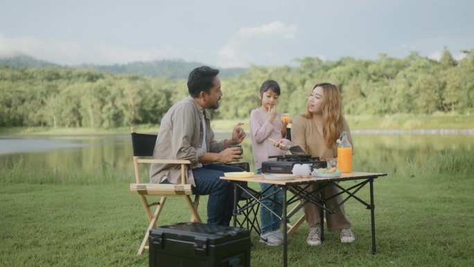 快乐的亚洲家庭，爸爸，妈妈和女儿在度假的草地上露营，享受食物和饮料。