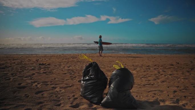 一个冲浪者在沙滩上走过，背景是一个冲浪板，前景是两个垃圾袋