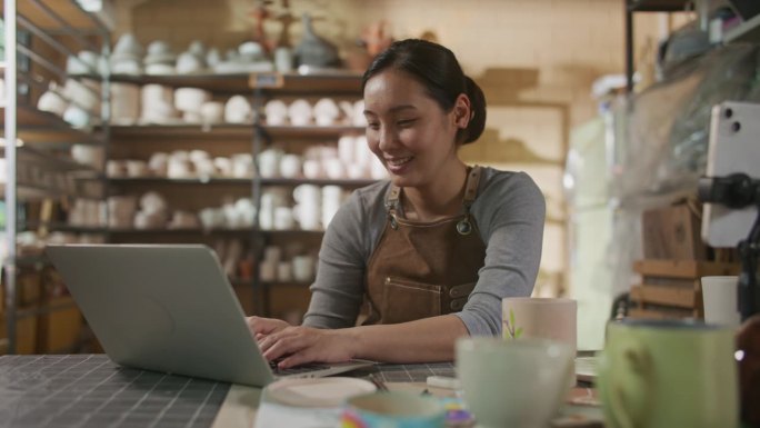 一名亚洲妇女在陶器工作室工作时使用笔记本电脑