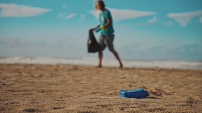一名成年男子在海滩捡垃圾，前景的沙滩上有一个蓝色的瓶子。塑料污染顺着河口流入海洋
