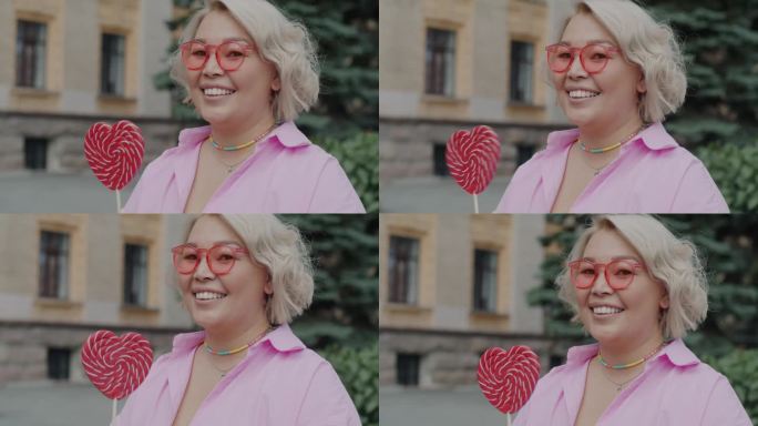 一名年轻漂亮的亚洲女子戴着粉红色的眼镜，手持心形糖果在城市户外的慢动作肖像