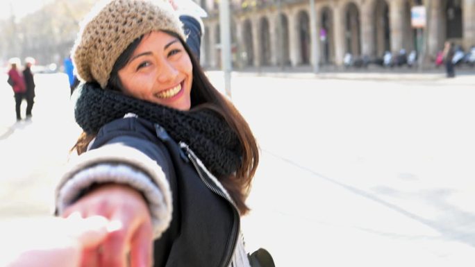 微笑的年轻女孩游客穿着外套，拿着宣传册，在城市街道上与男友手牵手，向镜头伸出手臂