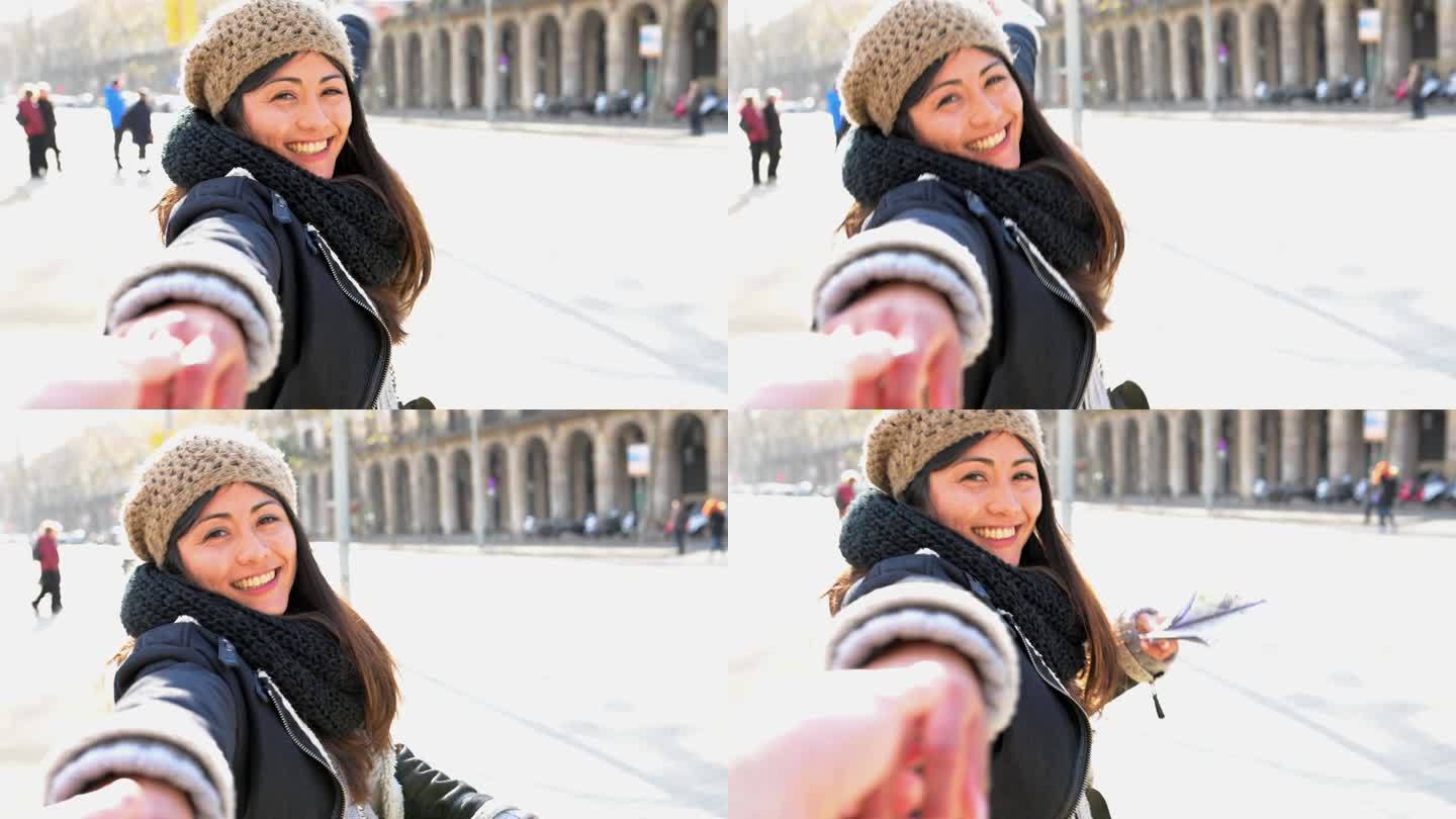 微笑的年轻女孩游客穿着外套，拿着宣传册，在城市街道上与男友手牵手，向镜头伸出手臂