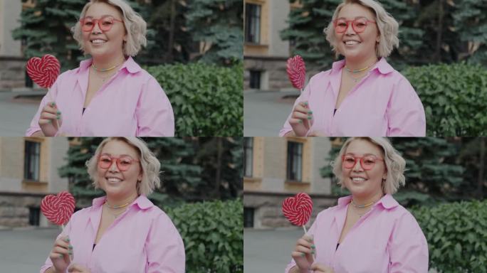 可爱的年轻女子的肖像，戴着粉红色的眼镜，站在城市街道的户外玩心形糖果