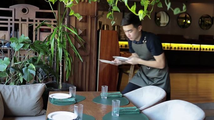 年轻的亚洲服务员在餐厅整理桌子。帅气的男员工穿着深色系带围裙在咖啡馆准备预定的桌子。食品和饮料概念。