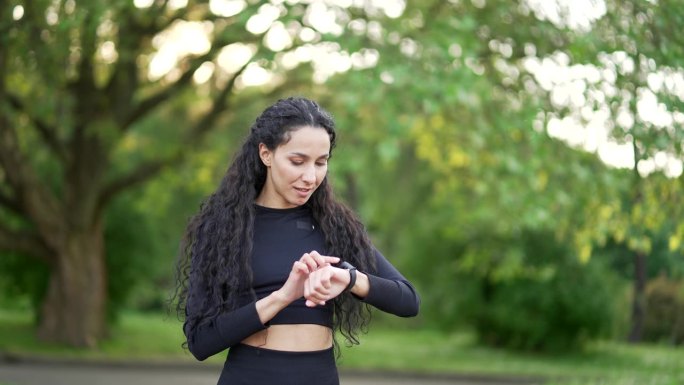 兴奋微笑的年轻女子跑步者看着智能手表在城市公园。女运动员使用检查健身追踪器