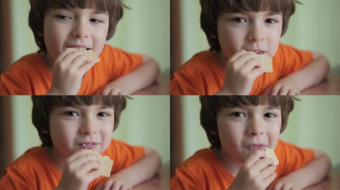 吃饼干的小男孩。看着镜头，微笑着。可爱的孩子饥饿吃饼干享受美味的治疗在家里的厨房。垃圾食品，不健康的