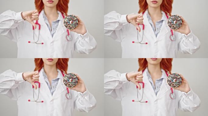 年轻的红发女医生拿着甜甜圈，在孤立的白色背景上做拇指向下的手势