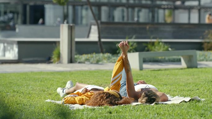女人，朋友和躺在草地上手牵手在公园暑假，休息或周末在城市。妇女们在野餐垫子上或阳光下的田野上放松，在