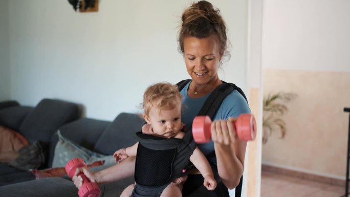 一名妇女在家里做健身运动，而婴儿则放在胸前