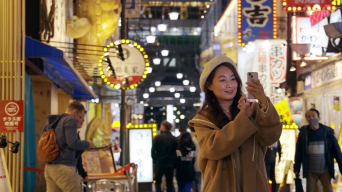 图为，一名亚洲女性在傍晚游览日本大阪的筑天阁塔。她用智能手机捕捉每一刻，记录她在热门景点的旅行经历。