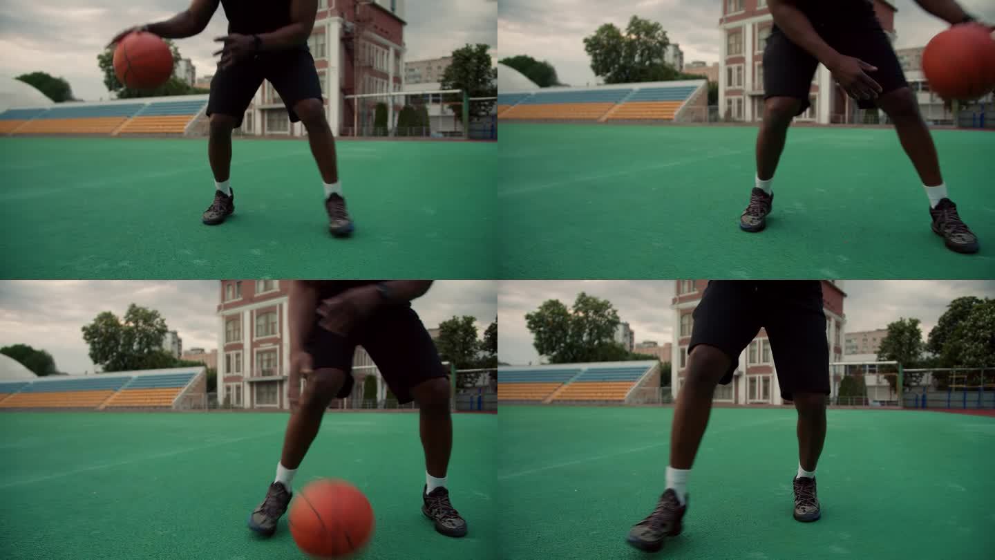 特写镜头，一名身穿黑色运动服的黑人篮球运动员将球击离地面，在灰色天空的衬托下对着城市中的体育场耍花招