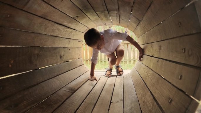 小男孩爬过操场上的木制隧道或管道。孩子们在户外玩耍，孩子们玩得开心，暑假和假期