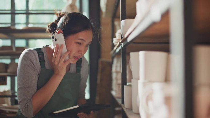 年轻的亚洲小企业主妇女用手机接收采购订单，并在她的工作室检查库存