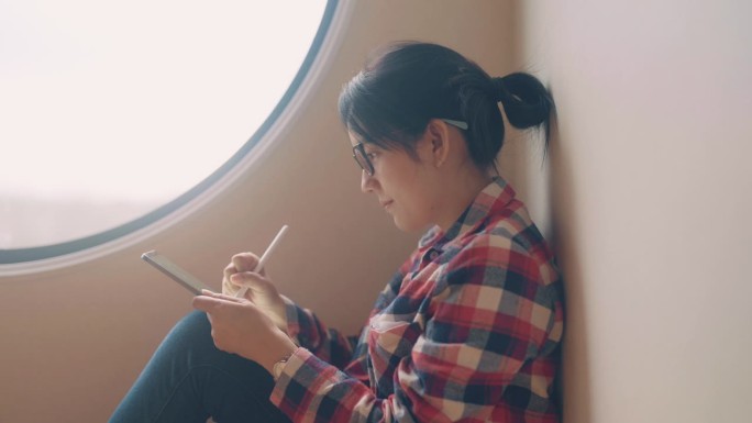 坐在客厅靠窗的年轻亚洲女子。她正在使用数字平板电脑，为项目创意写报告。