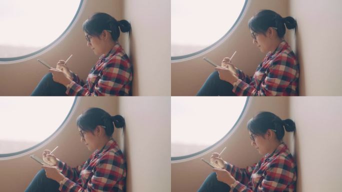 坐在客厅靠窗的年轻亚洲女子。她正在使用数字平板电脑，为项目创意写报告。